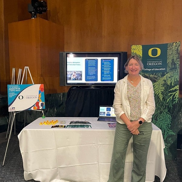 UO Professor Beth Stormshak in front of her presentation table at The Science of Addiction and Mental Health event. Screens and signs in the background are unreadable but showcase Professor Stormshak's work.